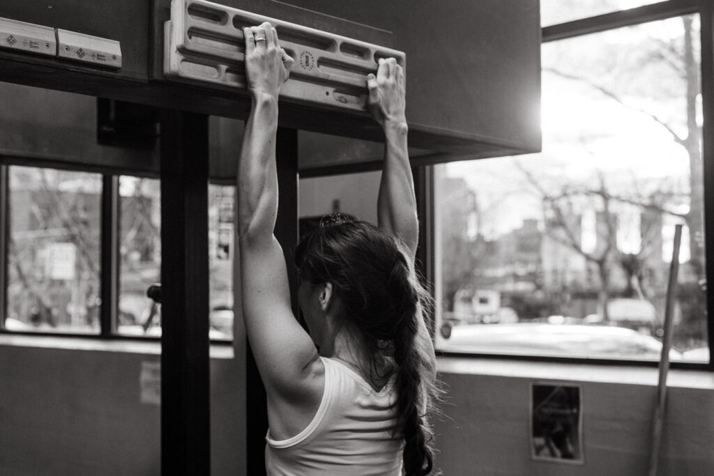 A woman with a braid hangs from a pull-up bar and climbing training board in a gym, viewed from behind. Sunlight streams through large windows, revealing an urban outdoor scene with trees and buildings.