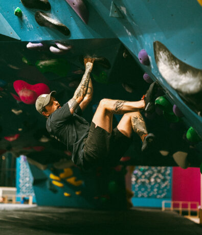 A person with tattoos and a cap climbs an indoor bouldering wall at a St Paul rock climbing gym, gripping holds on an overhanging blue surface. Their legs are bent as they hang close to the wall, focused on the next move.