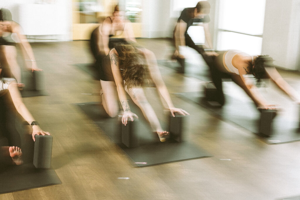 A group of people practicing Hot Fusion Yoga indoors, each on a mat using yoga blocks, captured in a motion-blurred effect that conveys movement and energy. The room has large windows and a wooden floor.