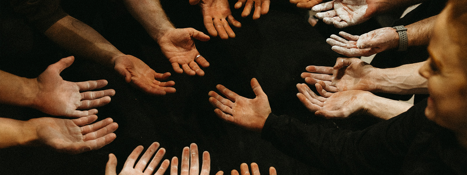 A group of people stand in a circle at Twin Cities Bouldering Project, extending their open, chalk-dusted hands toward the center over a dark surface.