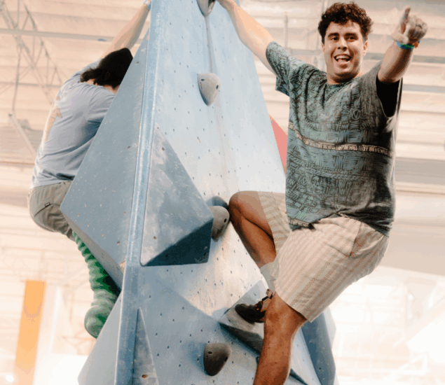 Two people are climbing an indoor blue geometric climbing wall. The man in front is smiling, pointing a finger, and appears happy and energized—perfect motivation for anyone considering a fall membership. The setting is bright and spacious.