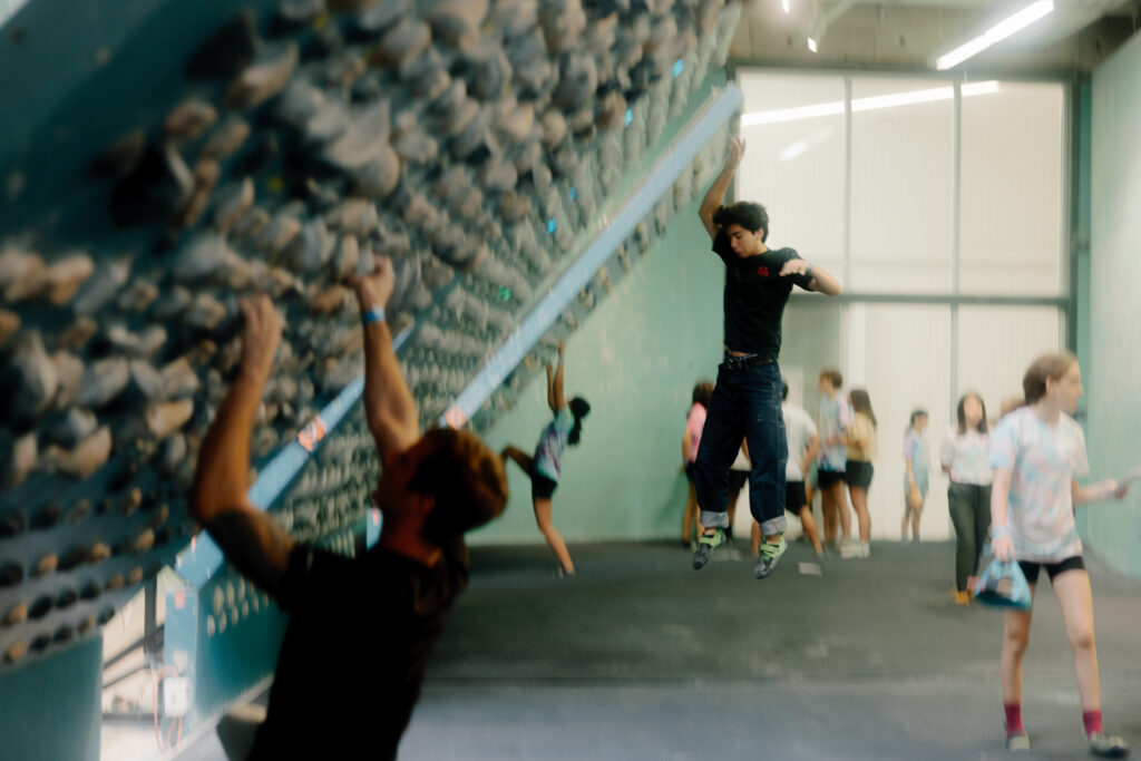 People climb on a steep indoor Kilter Board, with one person jumping off and others climbing or watching in the background. The wall is covered in holds, making it ideal for beginner to advanced climbers. The lively scene captures dynamic movement.