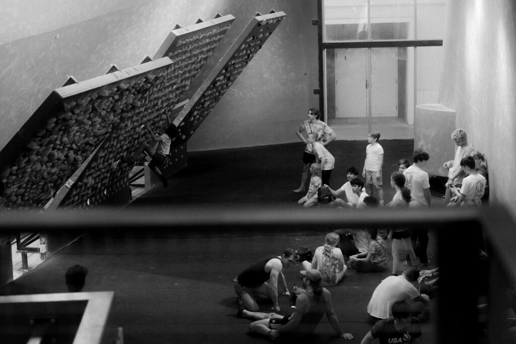 A group of people, some seated and some standing, watch a climber tackle an angled Kilter Board in a modern climbing gym. The scene captures the intensity of Kilter Board Climbing in striking black and white.