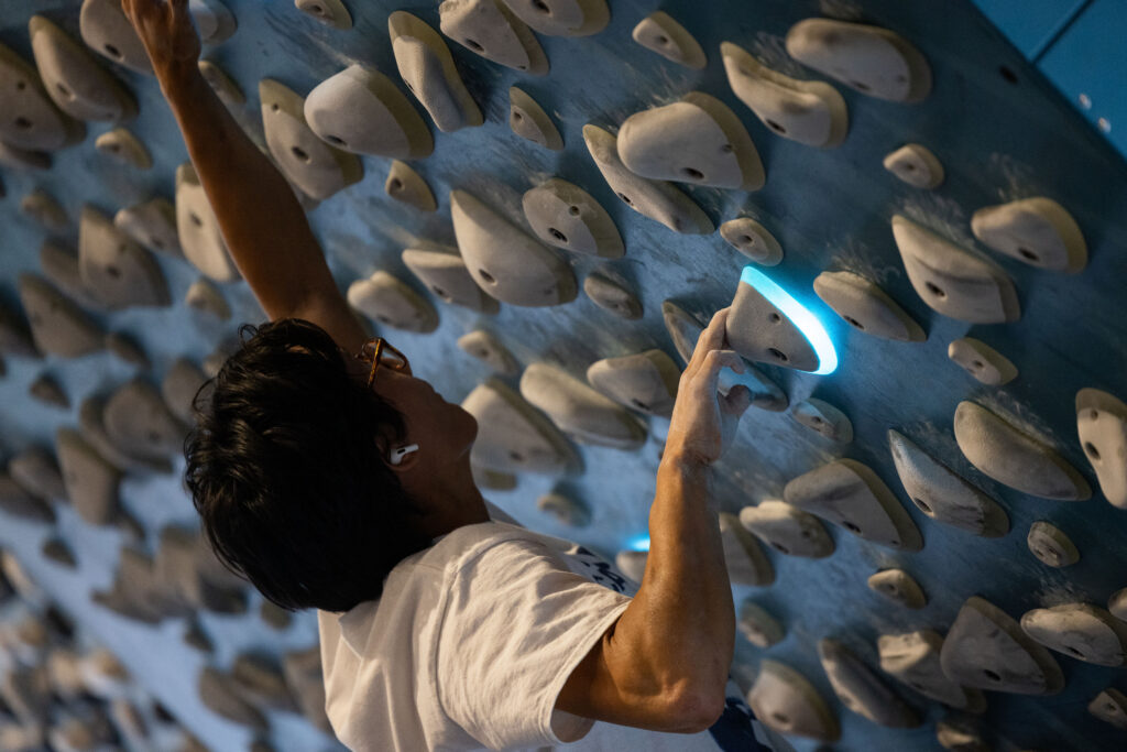 A person wearing glasses and a white t-shirt climbs a kilter board, reaching for illuminated holds among many gray handholds. The climber looks focused and determined, showcasing their passion for kilter board climbing.