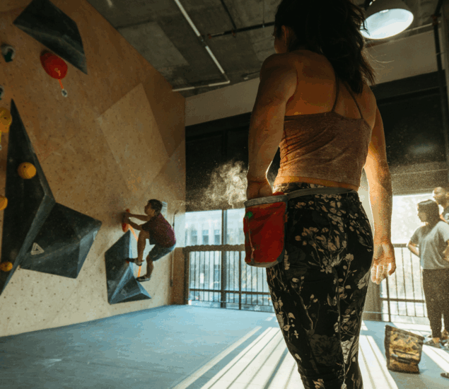 A woman in athletic wear stands in the foreground with chalk on her hand, watching another climber ascend an indoor bouldering wall. Sunlight streams through large windows as two people observe—perfect for New Year 2025 promos in All Markets.