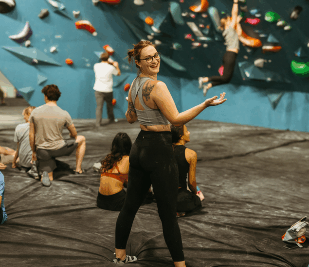 A woman in athletic wear smiles at the camera while standing on a padded floor at an indoor climbing gym, where others enjoy colorful holds. Get ready for New Year 2025 promos available across all markets!.