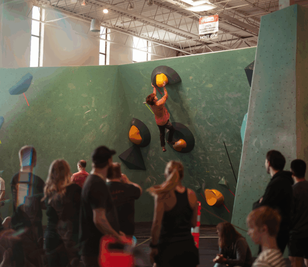 A climber hangs from a large yellow handhold on an indoor climbing wall in Minneapolis, watched by a group of people below during one of the gym's Skills Camps under high ceilings and bright lighting.