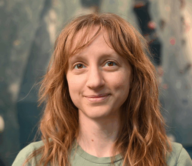 A woman with wavy, light brown hair and a green shirt smiles softly at the camera. The blurred blue and gray background hints at her calm confidence—perhaps inspired by Personal Climbing Coaching Austin.