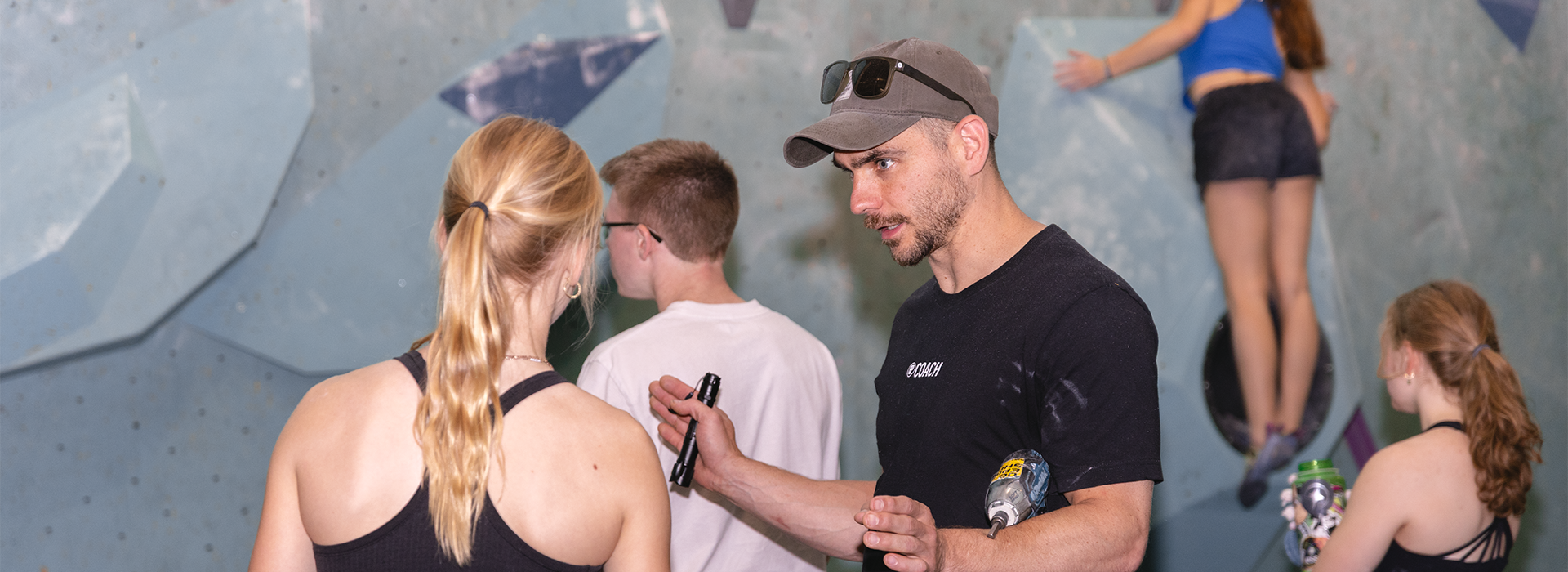 A man wearing sunglasses and holding a marker talks to a woman at an indoor climbing gym in Minneapolis, while two other people climb on the wall and one stands nearby, honing their skills as if training at specialized Skills Camps.