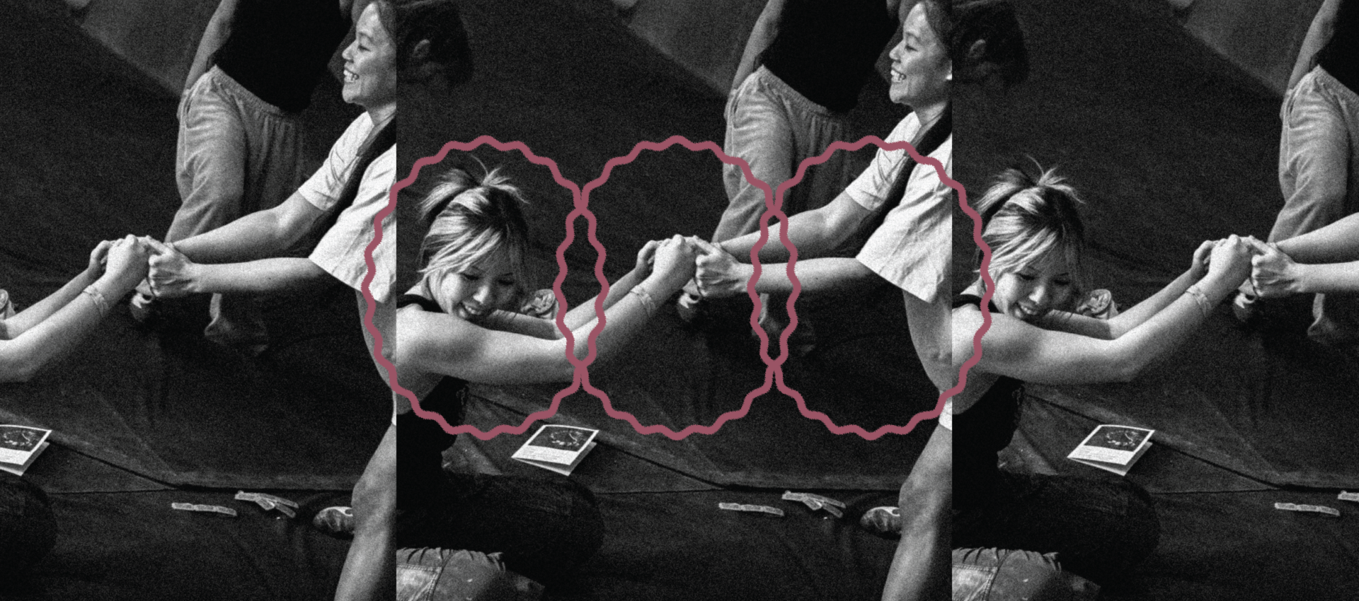 Black and white photo of two people at Seattle Boulderfest competition, sitting on mats, holding hands and pulling each other, with pink wavy lines circling their clasped hands. Other people and objects are partially visible in the background.