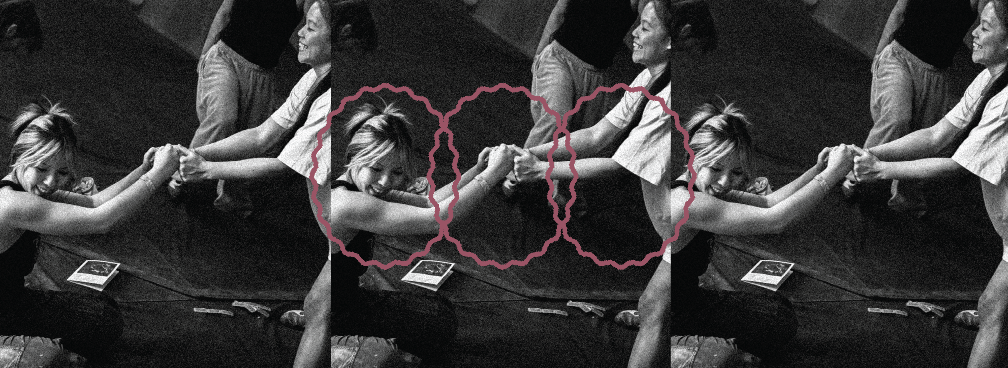 Black and white photo of two people at Seattle Boulderfest competition, sitting on mats, holding hands and pulling each other, with pink wavy lines circling their clasped hands. Other people and objects are partially visible in the background.