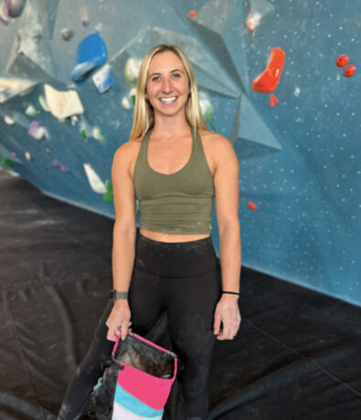 A woman in athletic wear stands and smiles in front of an indoor rock climbing wall, holding a colorful chalk bag—ready to make the most of her summer membership. The climbing wall behind her features various holds and geometric shapes.