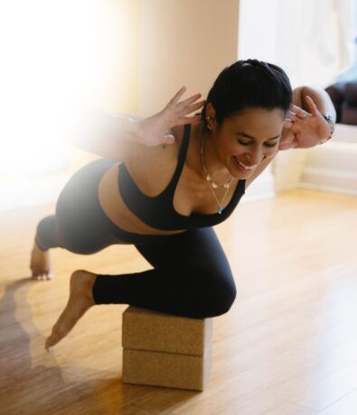 A woman in black athletic wear balances on yoga blocks in a yoga pose, smiling with hands by her head, during a personal training session on a wooden floor, sunlight streaming in from the left in DC.