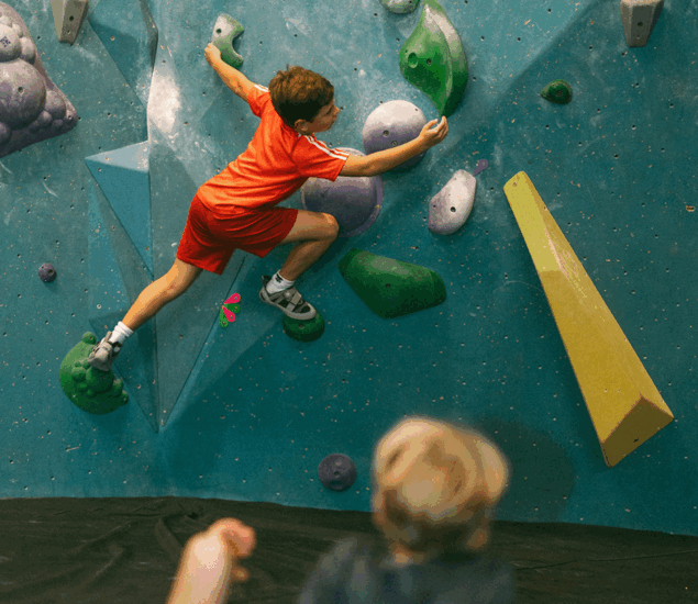A young boy in a red shirt and shorts climbs an indoor bouldering wall during Austin School Holiday Adventure Days, reaching for a green hold as another child watches. The wall features colorful holds and geometric shapes.