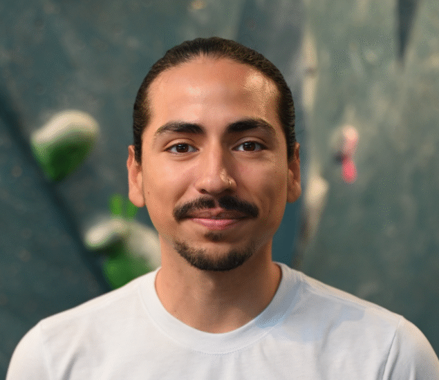 A man with long dark hair tied back, a mustache and goatee, wearing a light-colored T-shirt, smiles slightly while standing in front of a blurred indoor climbing wall, reflecting his passion for Personal Climbing Coaching Austin.