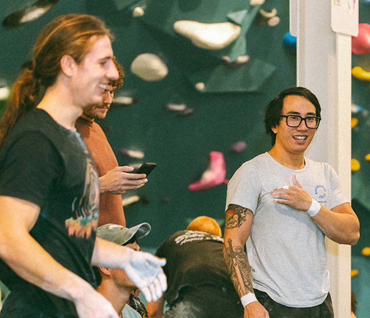 Two men with climbing chalk on their hands stand and smile in an indoor bouldering gym, surrounded by colorful holds and fellow climbers, capturing the spirit of Climb for Community.