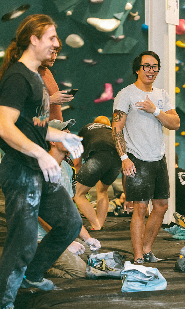 Two men with climbing chalk on their hands stand and smile in an indoor bouldering gym, surrounded by colorful holds and fellow climbers, capturing the spirit of Climb for Community.