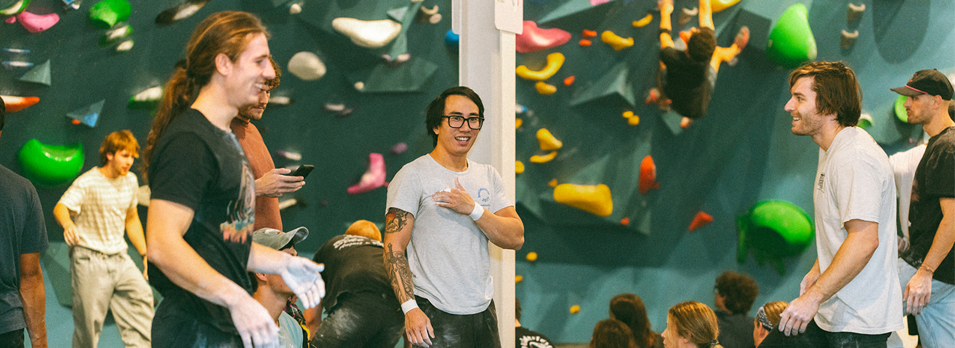 A group of people socialize and prepare for indoor rock climbing, with colorful holds in the background. Some are smiling and chalking their hands while others climb or watch, coming together to Climb for Community.