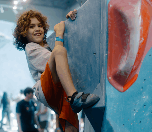 A smiling child with curly hair, wearing an orange shirt and climbing shoes, scales a blue indoor rock climbing wall with large red holds during Austin School Holiday Adventure Days. Other climbers are visible in the blurred background.