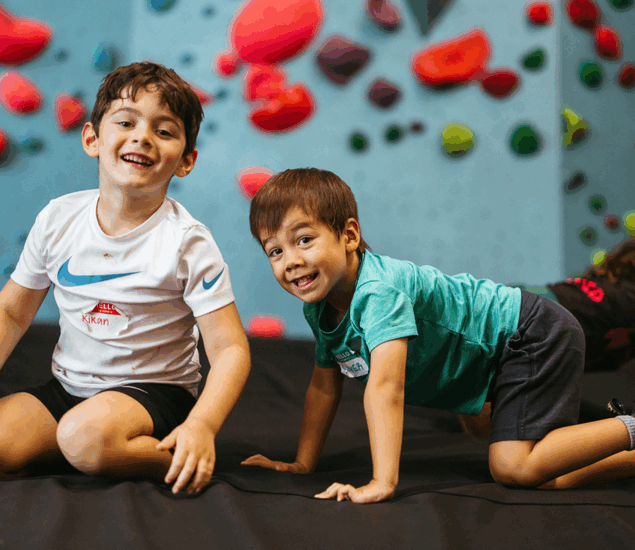 Two young boys smile and pose playfully on a padded floor at an indoor climbing gym during Austin School Holiday Adventure Days, with colorful climbing holds visible on the blue wall behind them.
