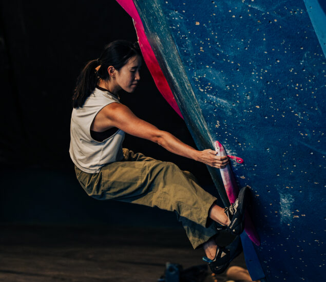 A woman wearing climbing shoes, a sleeveless shirt, and khaki pants grips a pink hold while bouldering on an indoor wall with blue and pink features during the Seattle Boulderfest competition.