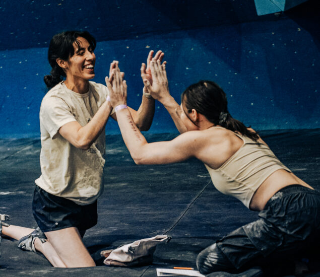 Two women kneel on a gym mat, smiling and giving each other a double high five after competing at the Seattle Boulderfest competition, with a blue climbing wall in the background.