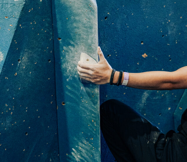A person’s hand gripping a white climbing hold on a blue indoor climbing wall at the Seattle Boulderfest competition, chalk marks visible. The climber sports several bracelets and a wristband; only their arm and part of their leg are seen.