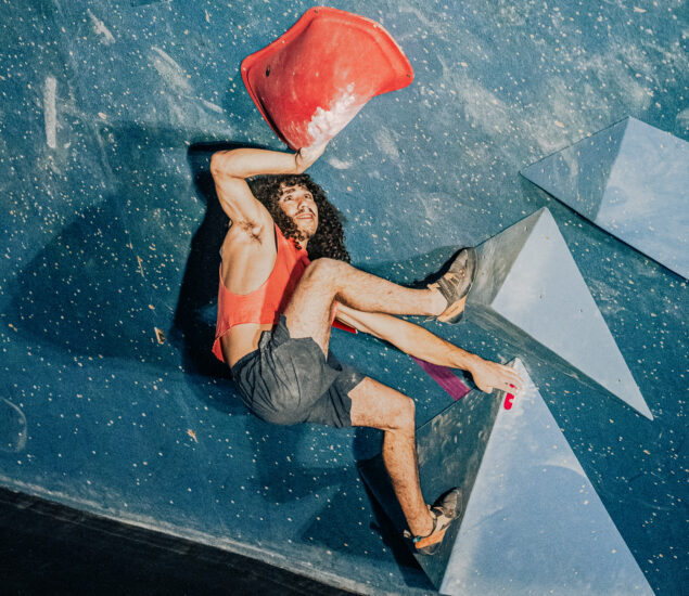 A person with curly hair wearing a red tank top and shorts climbs an indoor bouldering wall during the Seattle Boulderfest competition, gripping a large red hold and blue geometric volumes against a speckled blue background.
