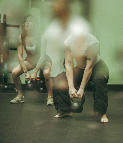 Two people are exercising indoors, each squatting with a kettlebell in hand—train smarter and climb stronger with these climbing exercises. The photo is softly focused; the front person is barefoot, while the one behind wears sneakers.