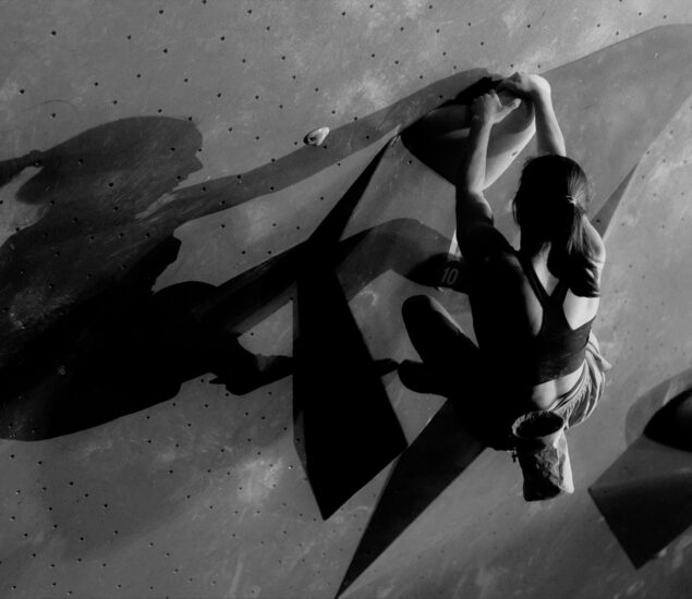 A climber in athletic wear clings to an indoor climbing wall, gripping holds with both hands and feet. Dramatic lighting casts deep shadows, emphasizing her strength and focus at Boulderfest 2026.