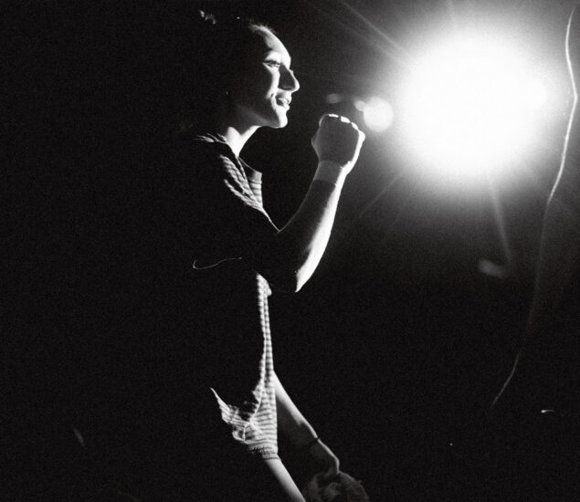 A woman, illuminated by a bright light source, stands smiling with her fist slightly raised. The black and white image, promoting Boulderfest 2026, creates dramatic contrasts and a vibrant sense of energy.