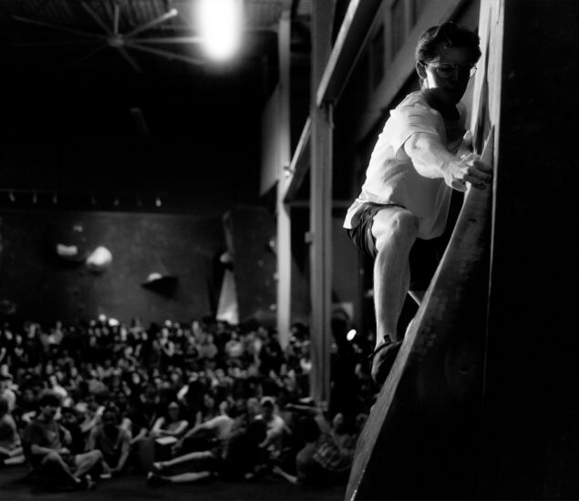 A person climbs an indoor bouldering wall in focus, while a large seated crowd watches in the blurred background. The scene is in black and white, with dramatic lighting highlighting the climber at Boulderfest 2026.