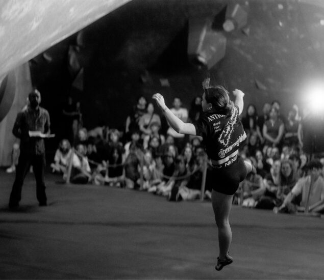 A climber leaps in mid-air during the Boulderfest 2026 indoor bouldering competition, with a crowd of spectators and a judge watching in the background. The scene is captured in black and white.