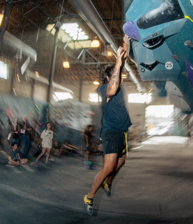 A person with an annual membership leaps toward a hold on an indoor climbing wall, mid-air, with one arm extended. Several people watch in the background of the brightly lit gym.