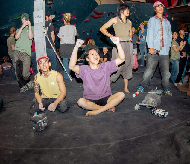 A group of people at an indoor climbing gym, with one person sitting cross-legged on the floor, raising their arms in excitement—perhaps celebrating a new route or an annual membership—while others and their climbing gear gather nearby.