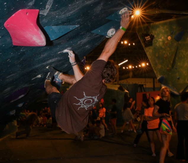 A man climbs an indoor bouldering wall, gripping handholds on an overhanging section during a competition. Spectators watch in the background under bright lights as the height fight unfolds. The climber wears a brown shirt and sports chalk on his hands.