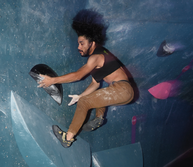 A person with long hair and a black crop top climbs an indoor bouldering wall, gripping a hold with both hands while balancing on blue holds with bent knees. The scene captures the intensity of a friendly community competition.