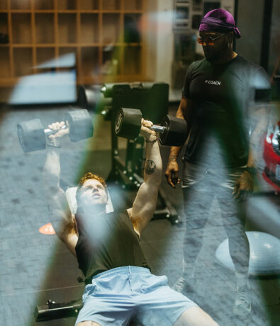 A man lies on a bench lifting dumbbells while another man, wearing a Coach shirt and a purple headband, supervises him in the gym—perfect motivation for anyone considering an annual membership.
