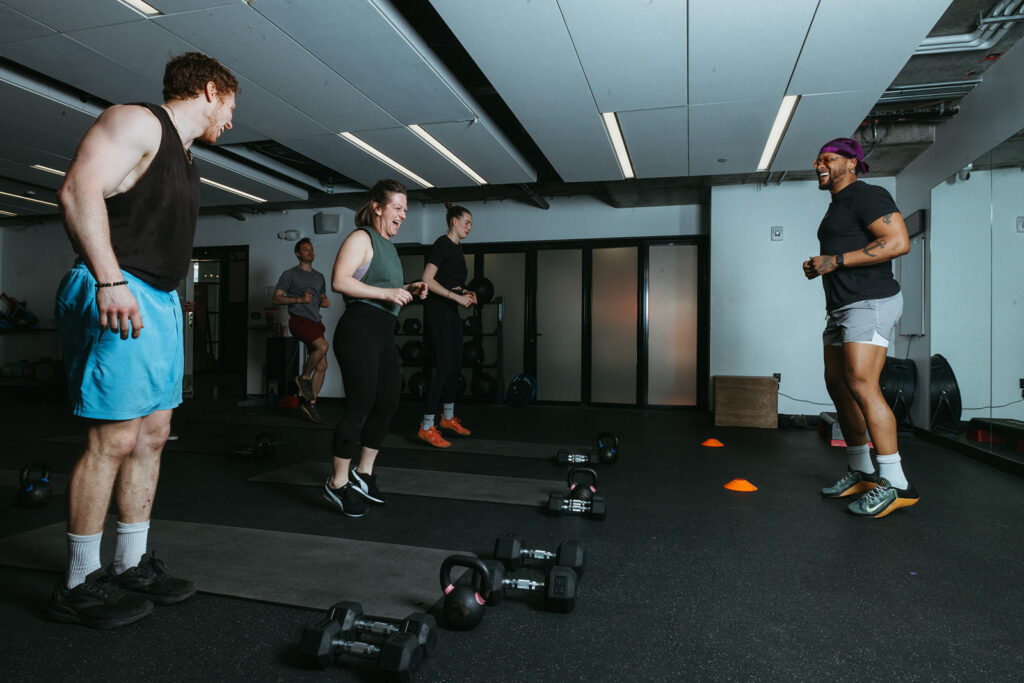 A group of people in athletic wear stand on exercise mats in a gym, listening and smiling at an instructor. Dumbbells and orange cones are on the floor around them as they prepare for climbing training to help them climb stronger.