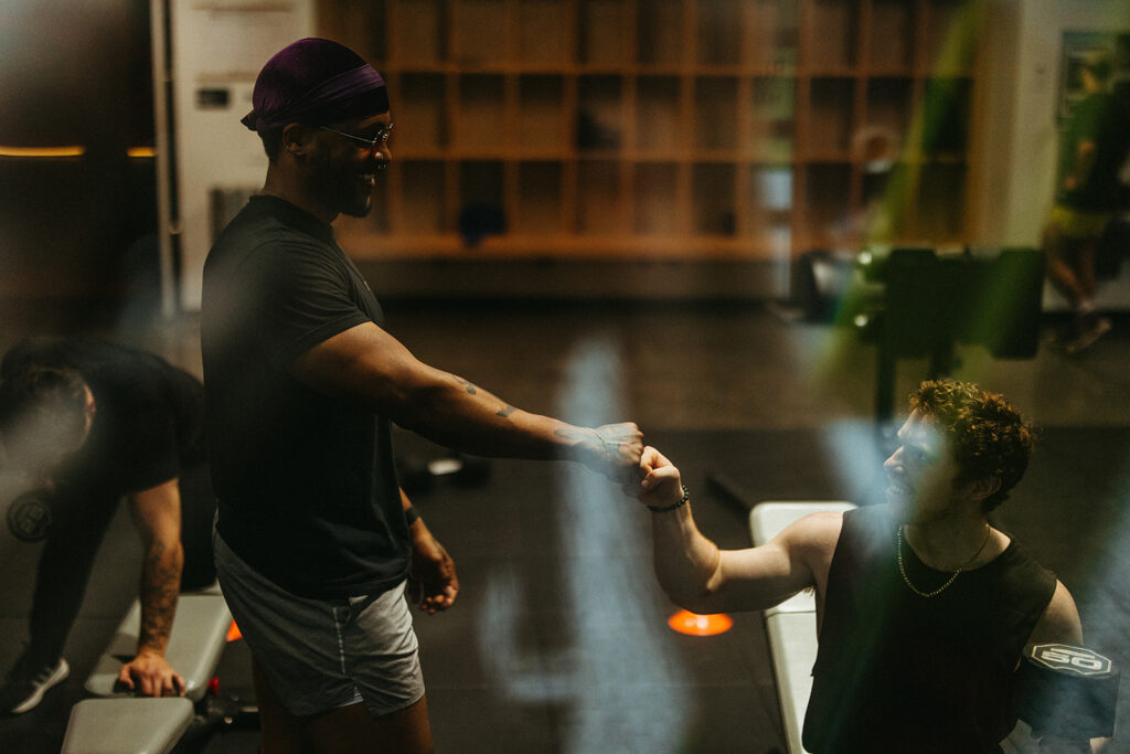 Two men at a gym fist bumping each other, one standing in shorts and a headwrap, the other sitting on a bench. Gym equipment and storage shelves are visible—perfect for those training for climbing or looking to climb stronger.