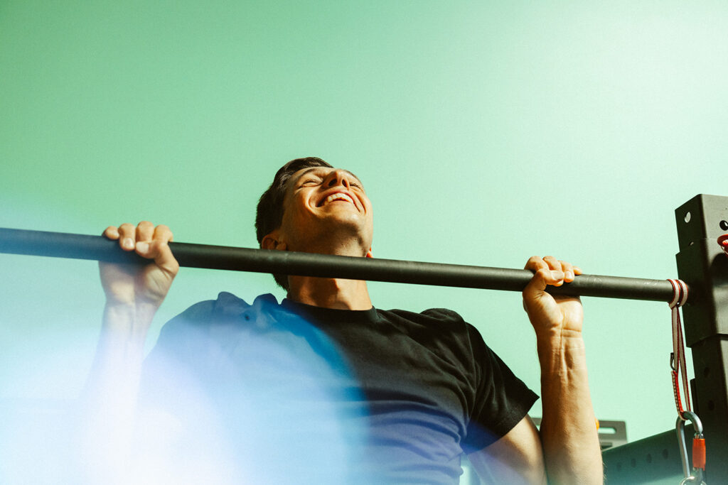 A man in a black t-shirt smiles as he does a pull-up on a horizontal bar against a light green background, with bright lighting and lens flare effects—demonstrating how to climb stronger with exercises for climbing.