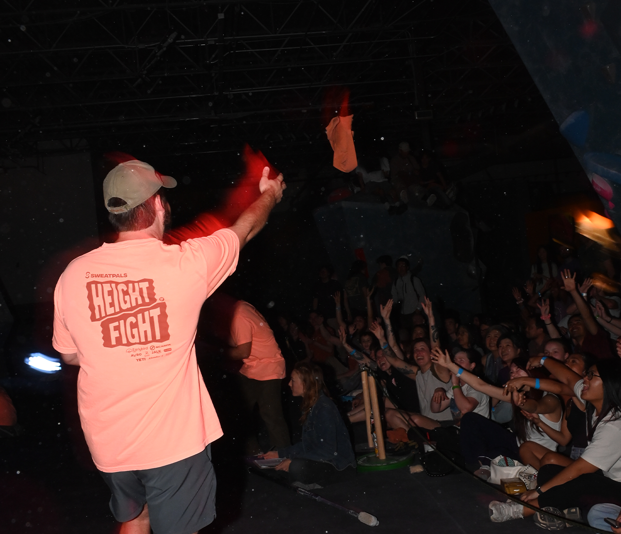 A man in a bright t-shirt throws an item into a crowd of excited people with raised hands at an indoor community event. The room is dimly lit, and everyone appears engaged in the spirited competition.