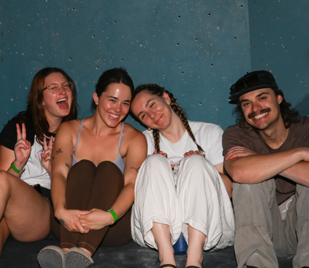 Four friends sit closely together on a couch against a teal wall, smiling and laughing after a fun height fight competition. One person makes a peace sign, while another rests her head on a friend’s shoulder. They appear relaxed and happy.
