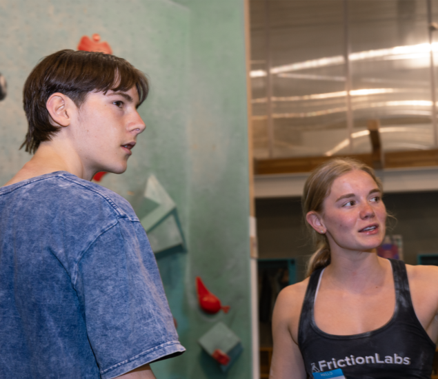 Two people stand indoors near a climbing wall in Austin, engaged in conversation. The person on the left wears a blue shirt, while the other sports a FrictionLabs tank top. Climbing holds are visible on the wall behind them, evoking summer camps.