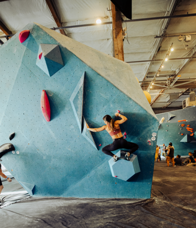 A woman in athletic clothing climbs a large blue indoor bouldering wall with geometric holds at Seattle Bouldering Project, where others watch and climb nearby—highlighting the vibrant community and membership benefits offered by the gym.