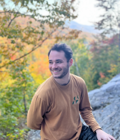 A young man with dark hair and a beard, wearing a brown long-sleeve shirt, smiles and looks to the side while sitting outdoors in Brooklyn, surrounded by colorful autumn foliage and a rocky landscape—perfect for personal coaching sessions.