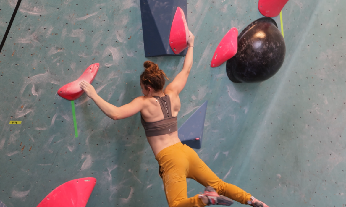 A woman wearing a sports bra and yellow pants climbs an indoor bouldering wall in the Twin Cities, reaching for pink holds with both hands and showing off her Skills Camps training as she balances expertly on lower holds.