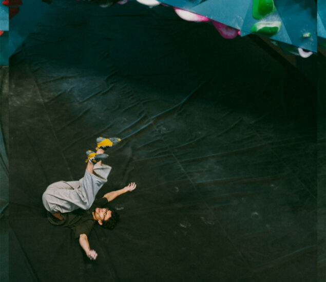 A person with a climbing gym membership, wearing climbing shoes and loose clothing, is falling onto a padded floor below an indoor climbing wall with colorful holds. Their arms and legs are extended as they land on their back.