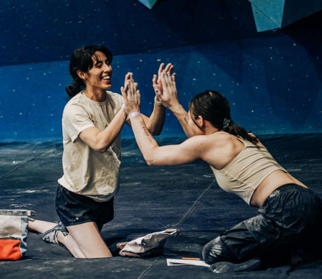 Two women kneel on the floor in front of an indoor climbing wall, smiling and giving each other a double high five, celebrating together. Climbing gear, a notebook, and info about Membership Benefits at Seattle Bouldering Project are on the ground nearby.