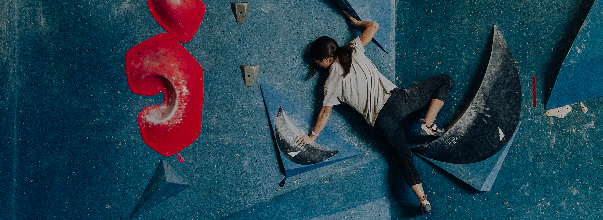 A woman wearing a white shirt and dark pants climbs indoors on a blue bouldering wall with large red and black holds, showcasing the exciting membership benefits at Seattle Bouldering Project.